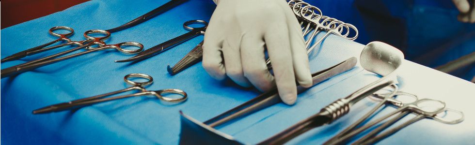 Die Hand eines Chirurgen greift nach einer Pinzette aus dem chirurgischen Nahtset, welches auf dem Instrumententisch liegt
