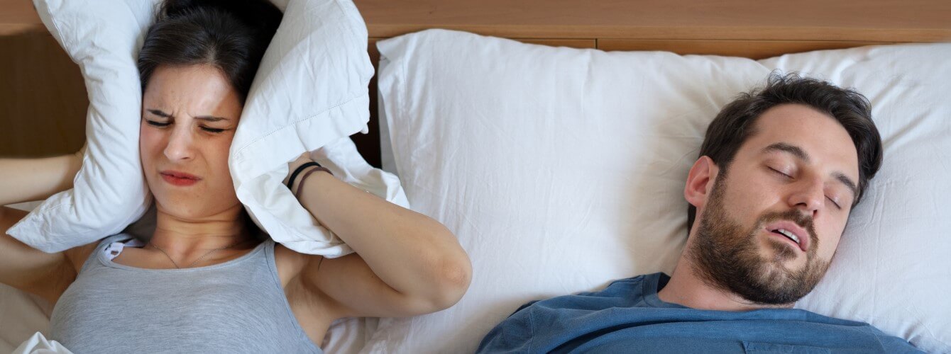 Eine Frau hält sich mit einem Kissen die Ohren zu, weil der Mann neben ihr im Bett aufgrund von Schlafapnoe schnarcht